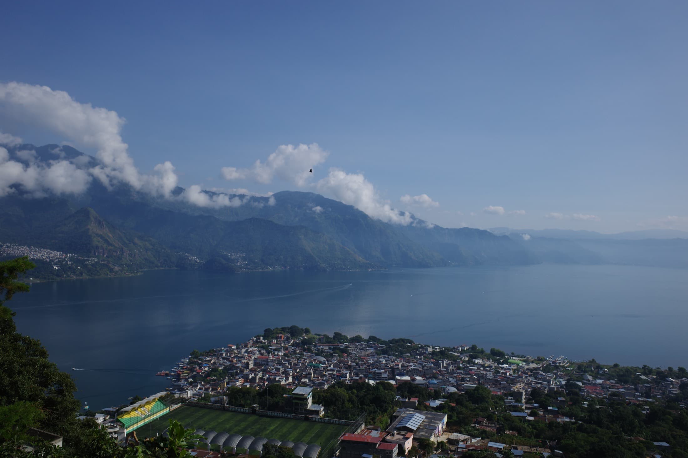 Lake Atitlán from above — the deep-blue caldera lake ringed by volcanoes, the town of San Pedro and its football pitch spilling down to the shore, a hawk riding the thermals.