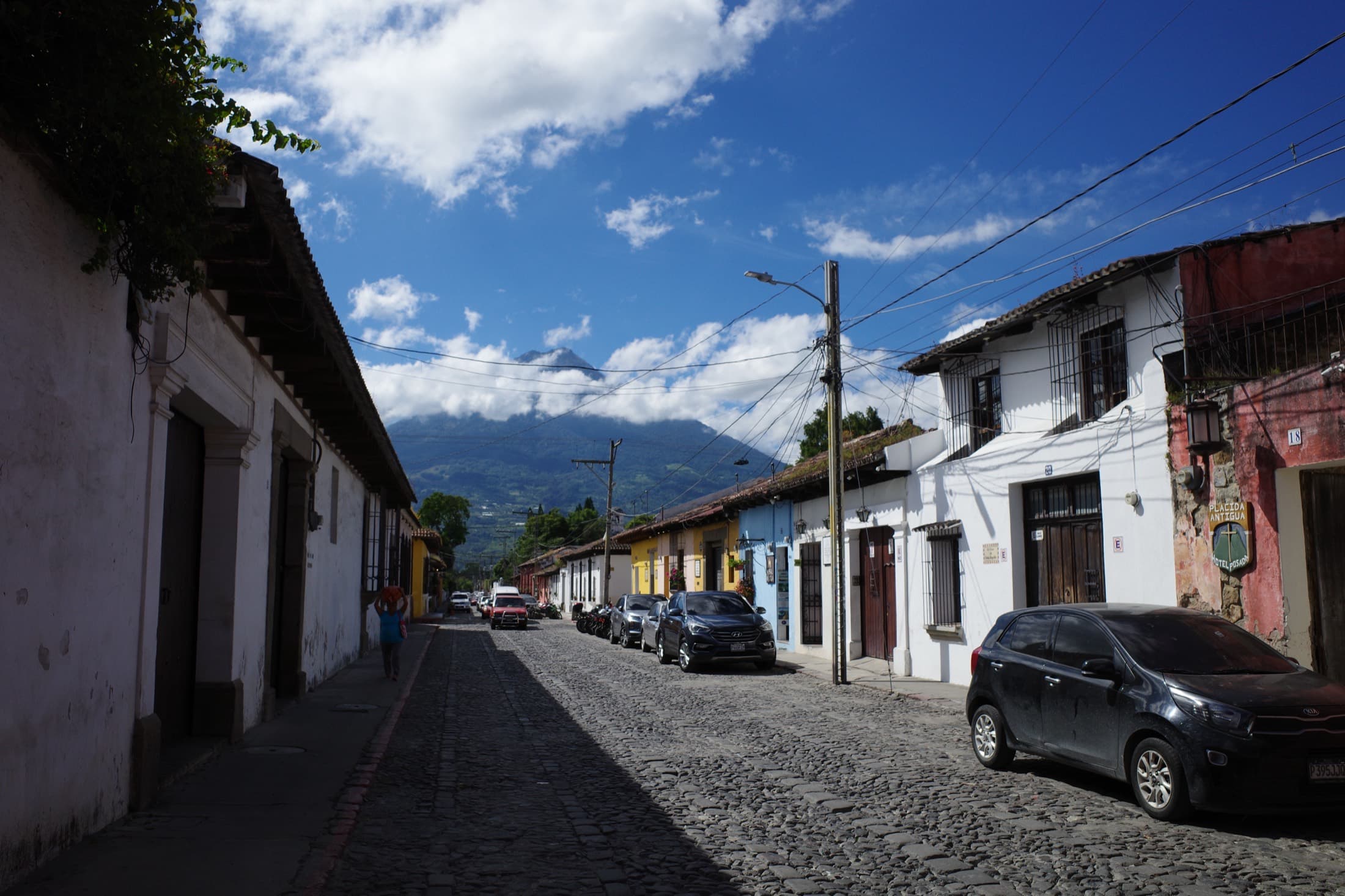 A cobblestone street in Antigua, Guatemala — pastel colonial houses and parked cars lining the way, Volcán de Agua rising through the clouds at the end.