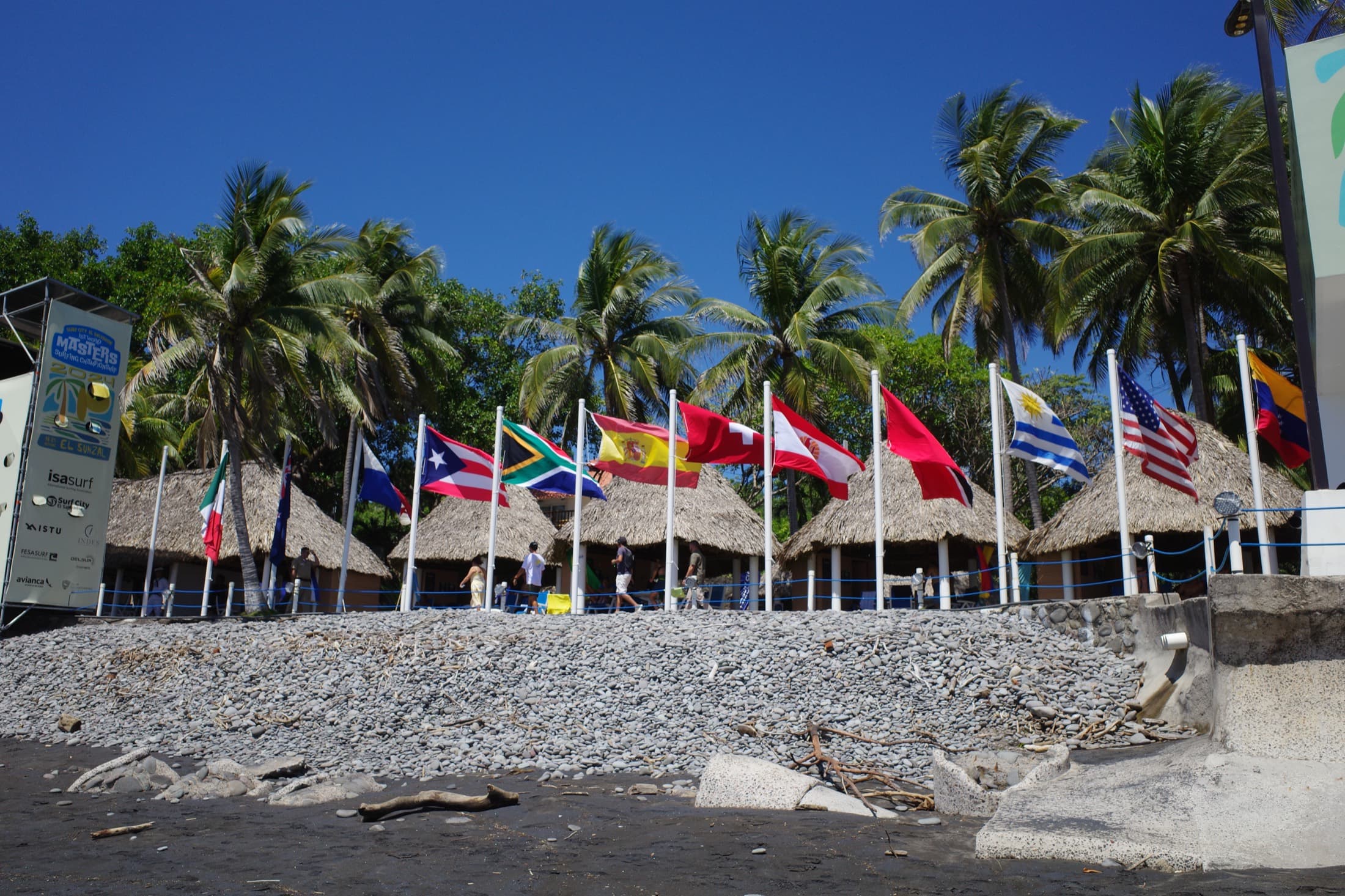 Flags of the surfing nations lined up on the black-sand beach at El Sunzal, El Salvador, during an ISA world surf event — palms and thatched ranchos behind.