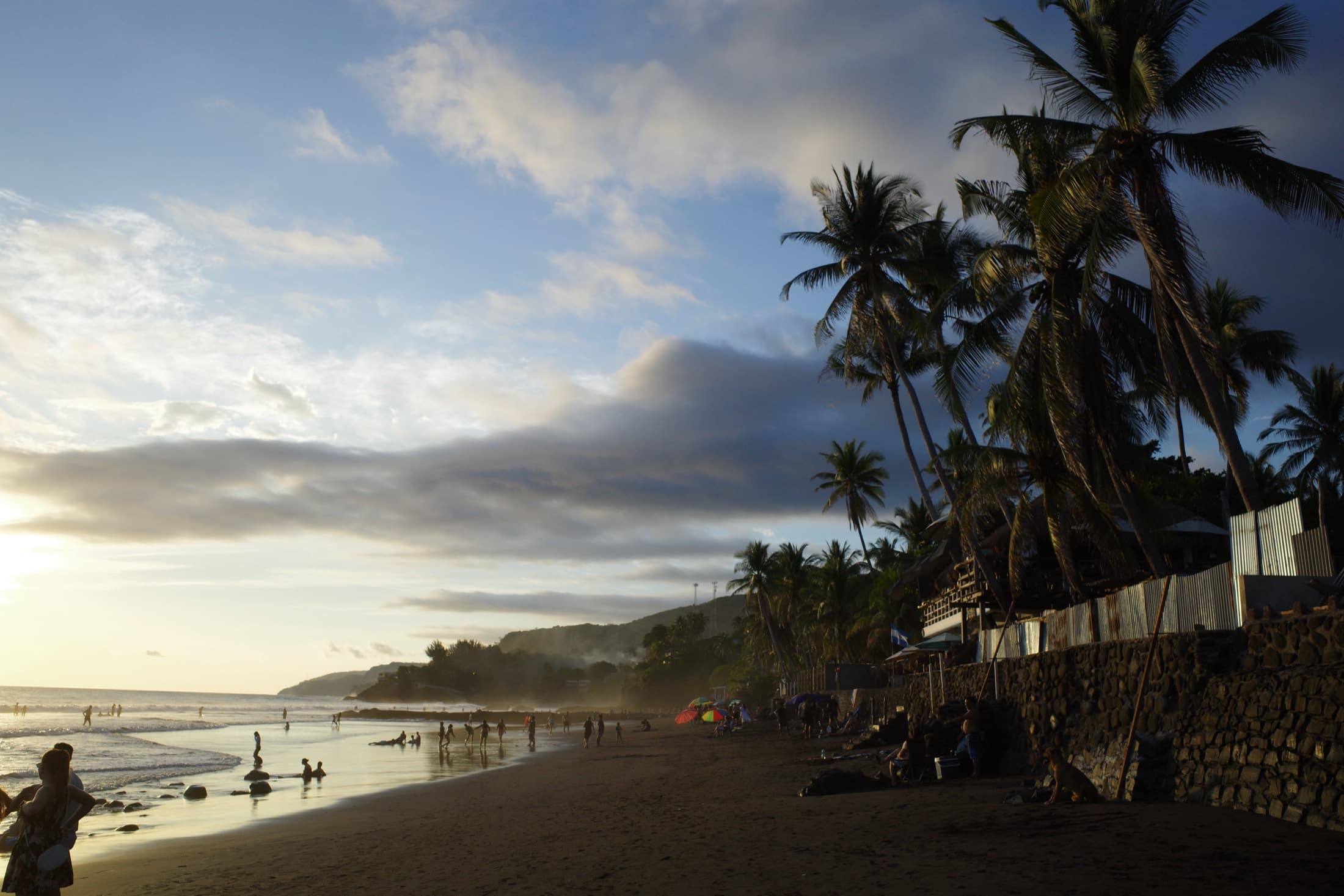 Sunset on the surf beach at El Tunco, El Salvador — palms leaning over the sand, families in the shallows, the point break misting in the distance.