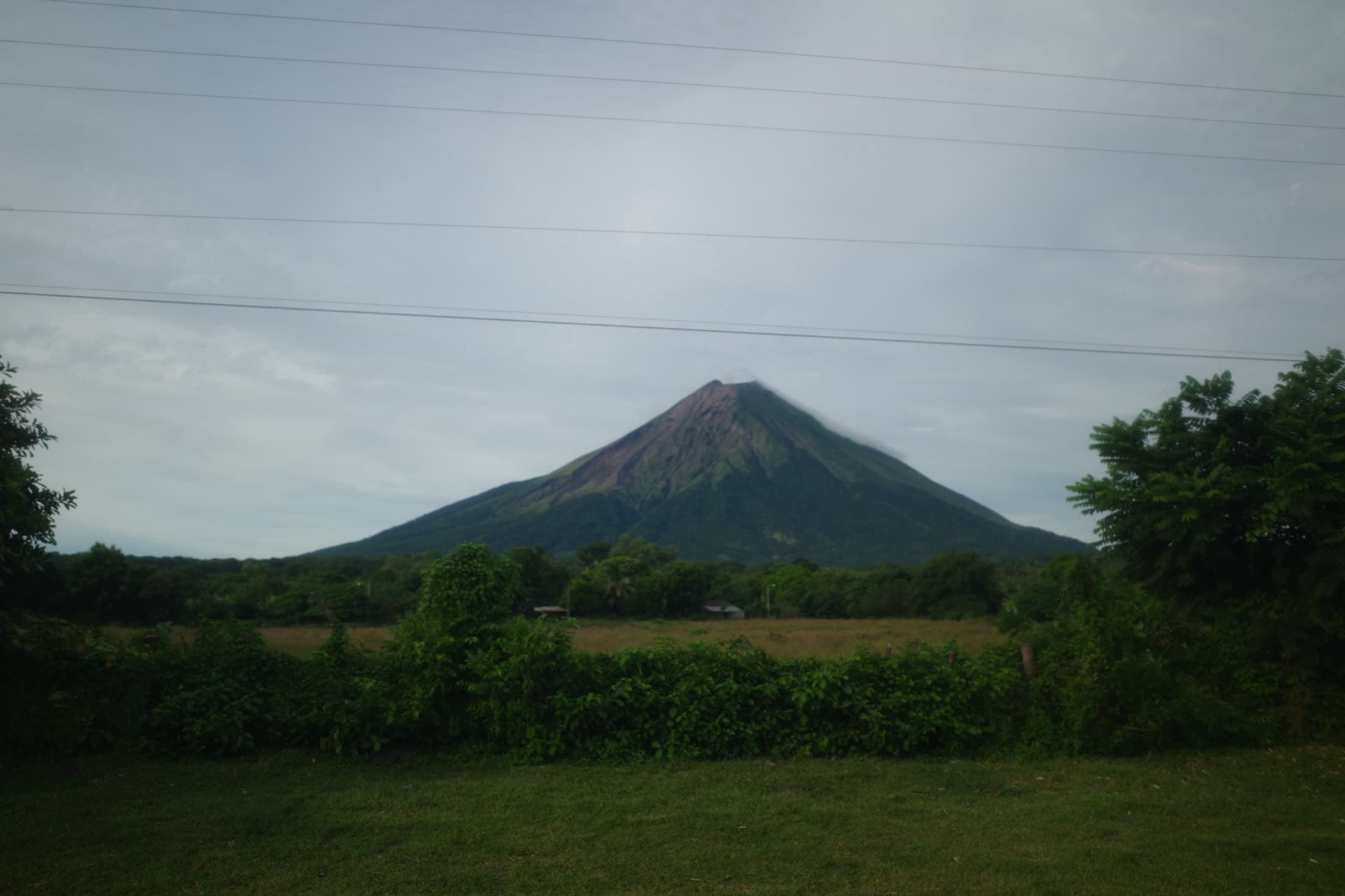 Volcán Concepción rising over the fields of Ometepe island, Nicaragua — a near-perfect cone under a soft grey sky, power lines strung across the foreground.