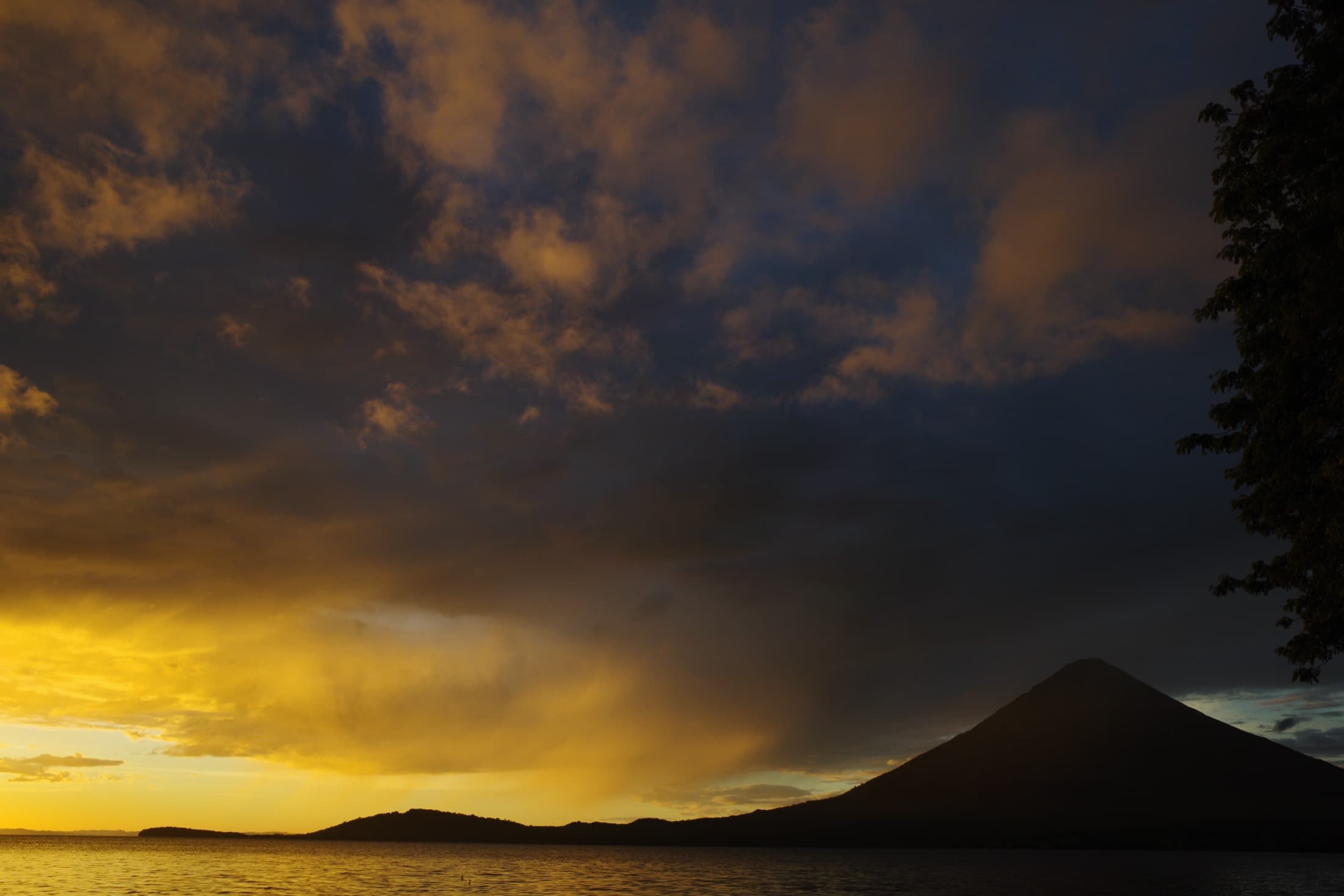 A dramatic golden sunset over a Nicaraguan lake — storm light breaking through the clouds behind a volcano silhouette.