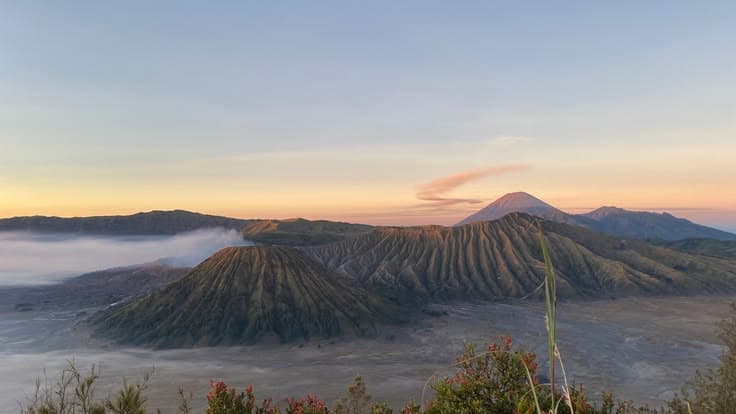 Mount Bromo at sunrise in East Java — the smoking caldera and its neighbouring cones rising from a sea of sand and mist, the peak of Semeru catching the first pink light behind.