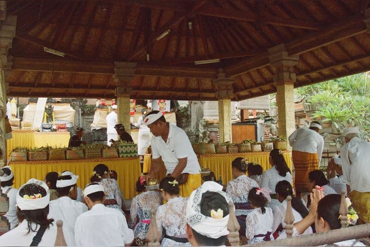 A Balinese temple ceremony under a carved wooden bale — rows of worshippers in white lace and headscarves seated with offerings, a man in a white udeng handing out blessings by a gold-draped shrine.