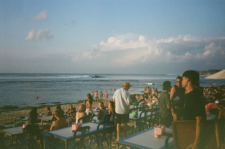 A cliff-top warung above Bingin beach in Bali at golden hour — a crowd of surfers and travellers at plastic tables watching a clean reef break peel in the distance.