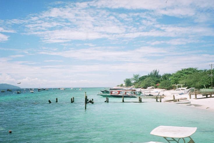 The turquoise shallows of Gili Trawangan — wooden jetty posts, local boats moored off a white-sand beach, a wispy blue sky over the calm water.
