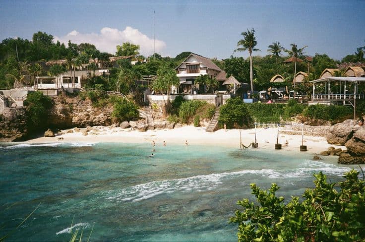 A hidden cove on Nusa Lembongan — cliffside villas and warungs above a clear turquoise bay, swimmers in the water, a beach volleyball net and hammocks on the sand.