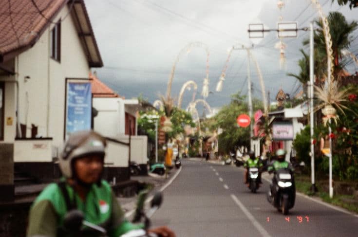 A Balinese village street decked with towering penjor bamboo poles for Galungan — a green-jacketed Gojek driver on a scooter, misty mountains and a red stop sign down the road.