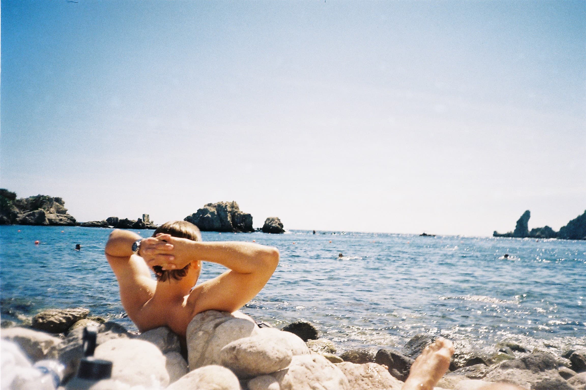 An Italian beach in afternoon light — a sunbather on the warm rocks, rugged islets in the bay.