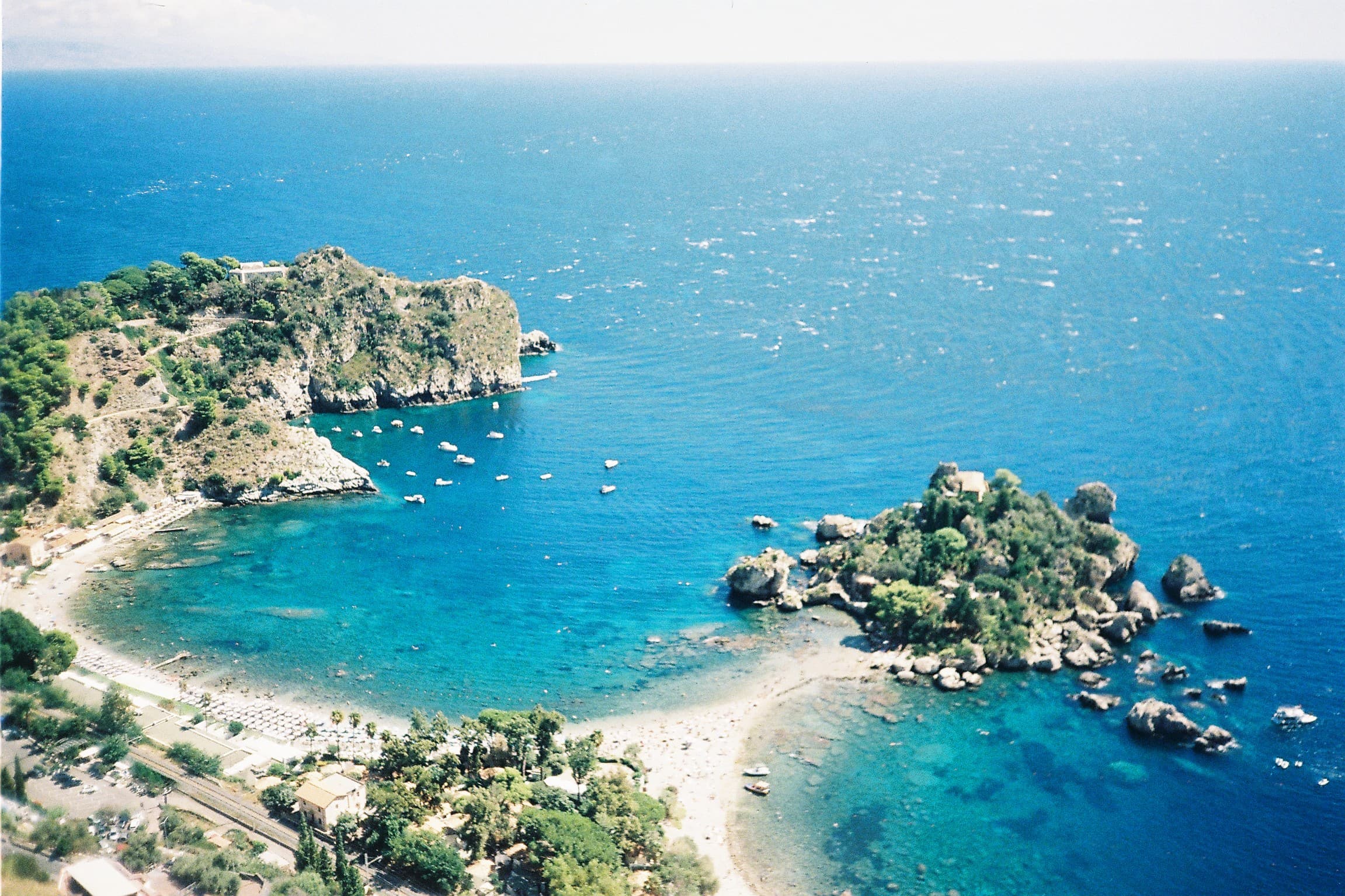 Isola Bella in Taormina from above — the islet connected to the mainland by a sand bar, deep blue Ionian Sea around it.