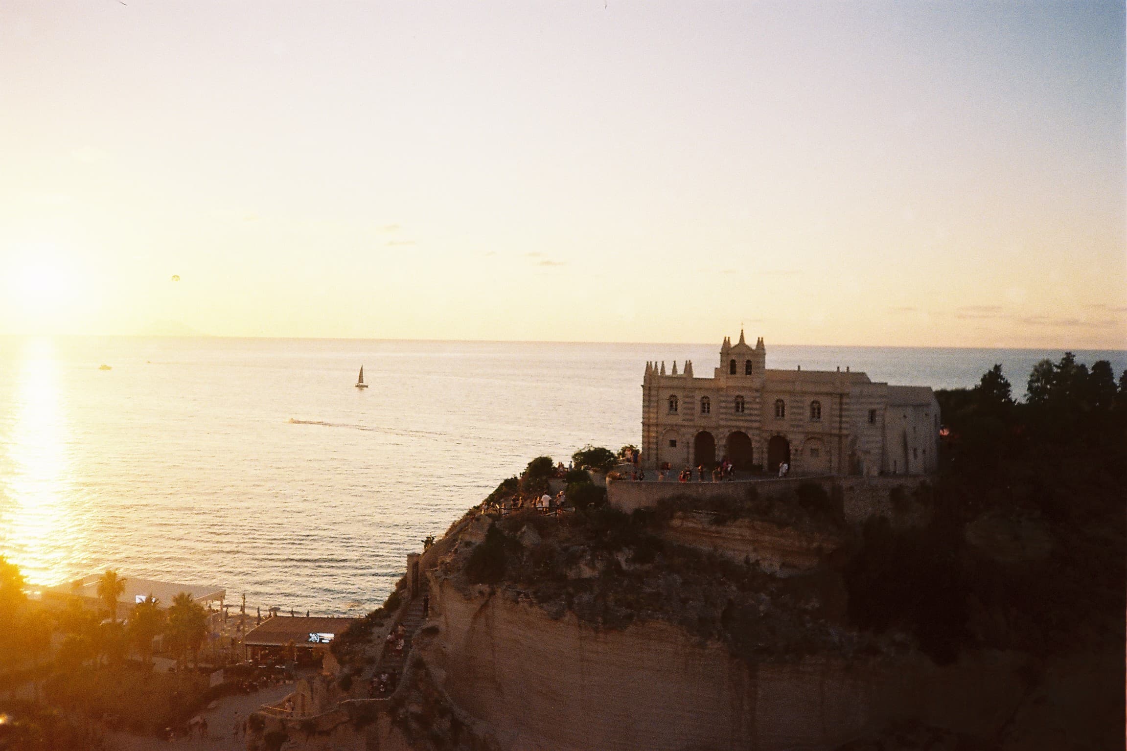 Santa Maria dell'Isola at Tropea at sunset — the clifftop monastery silhouetted, sailboat on the horizon, soft Calabrian light.