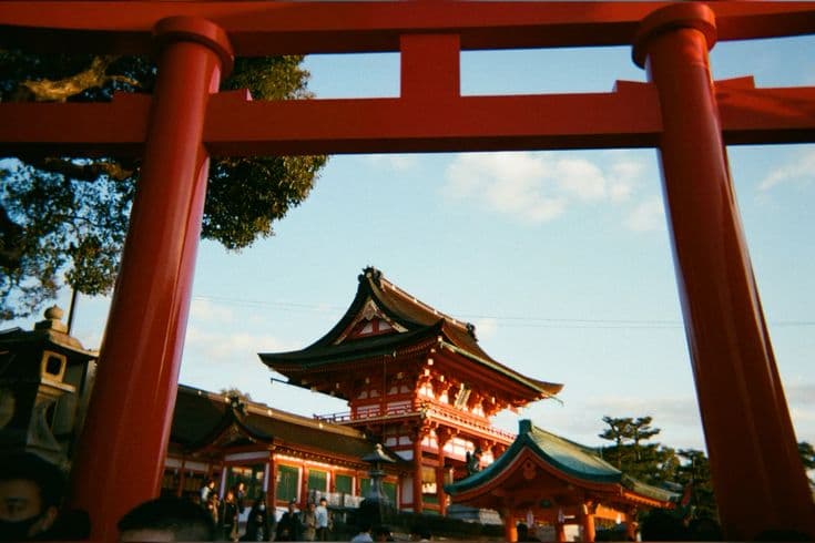 Fushimi Inari shrine in Kyoto framed through a giant vermilion torii gate — the ornate main hall glowing at dusk, worshippers gathered below.