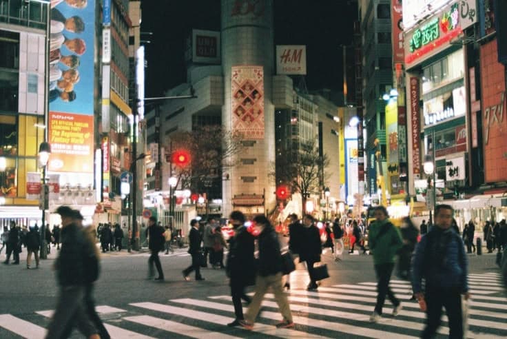 Shibuya Crossing at night — crowds streaming across the scramble under a wall of neon and billboards, Uniqlo and H&M signs lighting the Tokyo dark.
