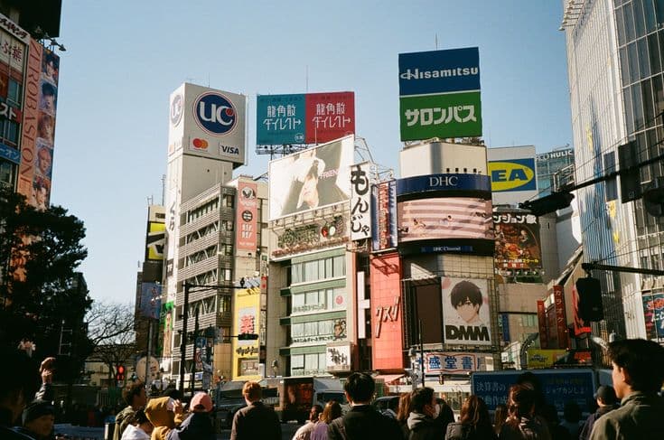 A Tokyo street by day stacked with billboards and Japanese signage — a crowd waiting at the lights beneath the layered advertising of Shibuya.