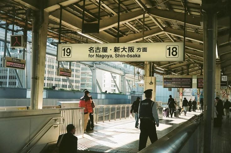 A Shinkansen platform — the overhead sign reading 'for Nagoya, Shin-Osaka', a station guard and travellers with cases waiting for the bullet train under the steel canopy.