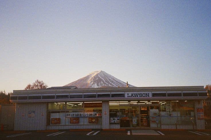 A roadside Lawson convenience store at dawn with Mount Fuji rising directly behind it — the quiet, everyday Japan that stops you in your tracks.