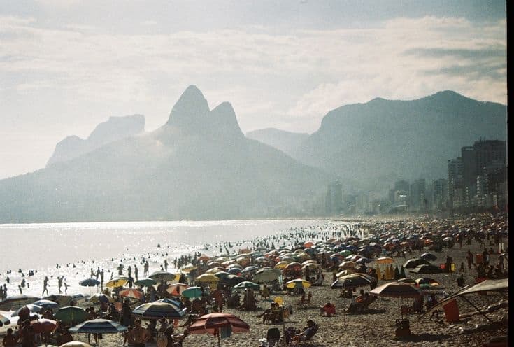 Ipanema beach in Rio de Janeiro on a hazy golden afternoon — a sea of colourful umbrellas and swimmers, the Dois Irmãos (Two Brothers) peaks rising behind the bay.