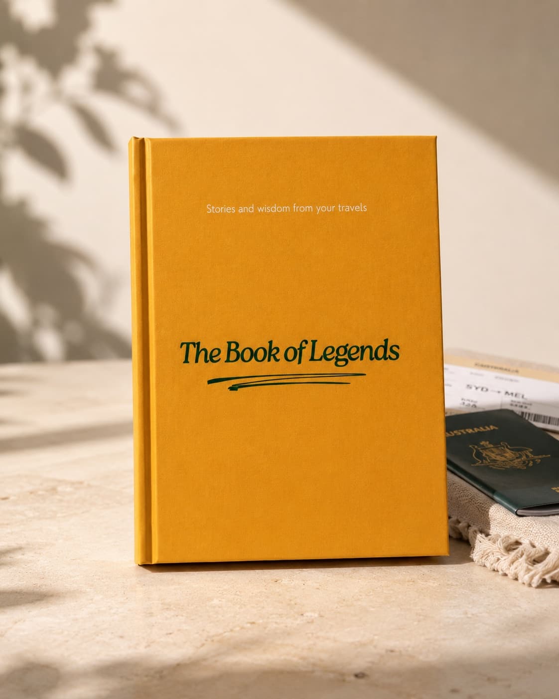 Front cover of The Book of Legends — orange hardcover with 'Stories and wisdom from your travels' above the title in deep green, photographed on warm stone next to an Australian passport and a SYD→MEL boarding pass