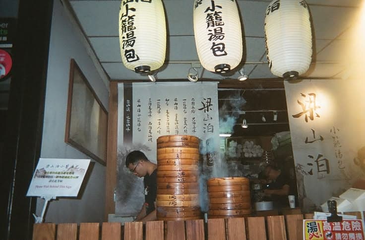Stacked bamboo steamer baskets billowing steam at a xiaolongbao (soup dumpling) shop, big white paper lanterns lettered 梁山泊 小籠湯包 hanging above the counter, cooks at work behind.