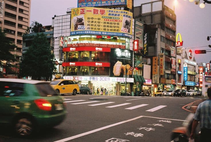 A neon-lit Taichung street corner at dusk — layered Mandarin shop signs stacked up a building, a yellow taxi and a car blurring through the crossing.