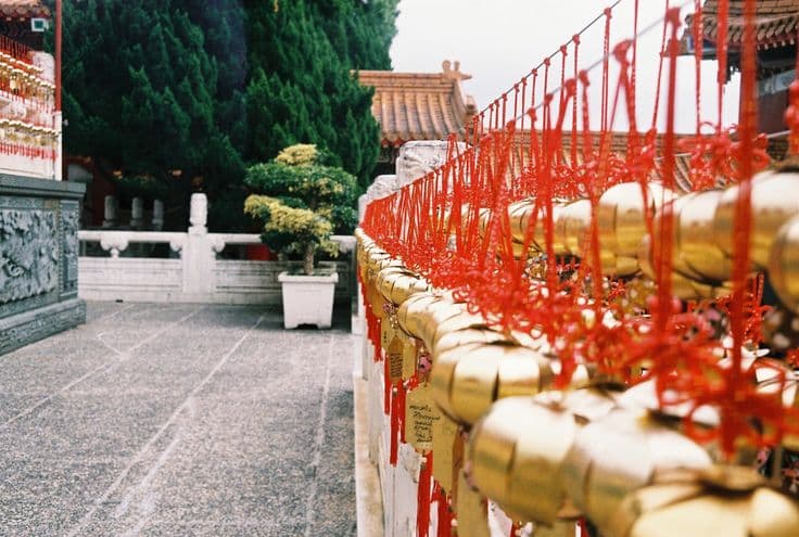 Rows of gold prayer bells strung with red ribbons along a balustrade at Wenwu Temple, Sun Moon Lake — ornate temple roofs and manicured pines beyond.