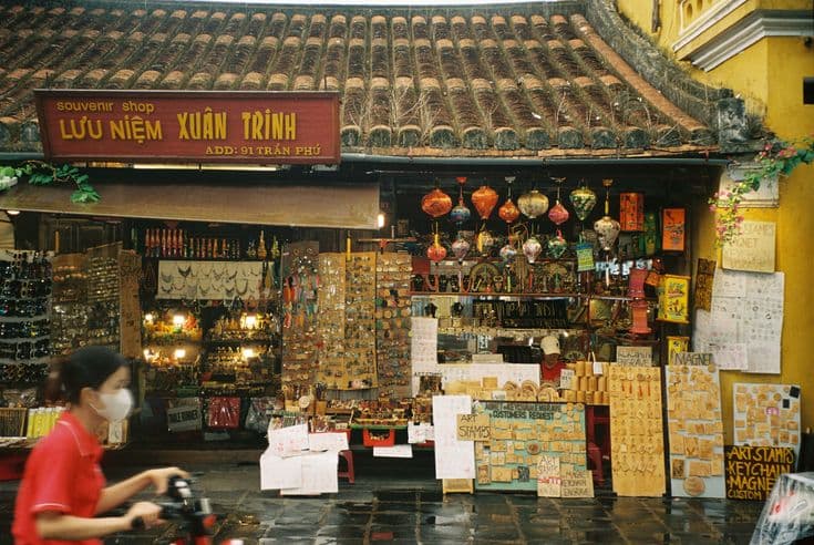A Hoi An old-town souvenir shop under a tiled roof — silk lanterns strung across the front, racks of art stamps and trinkets, a woman on a motorbike passing in red.