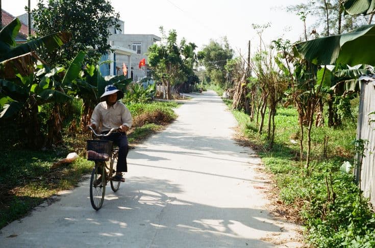 A man in a bucket hat cycling down a quiet concrete village lane near Hoi An — banana palms and greenery either side, a Vietnamese flag on a house, hard midday shadows.