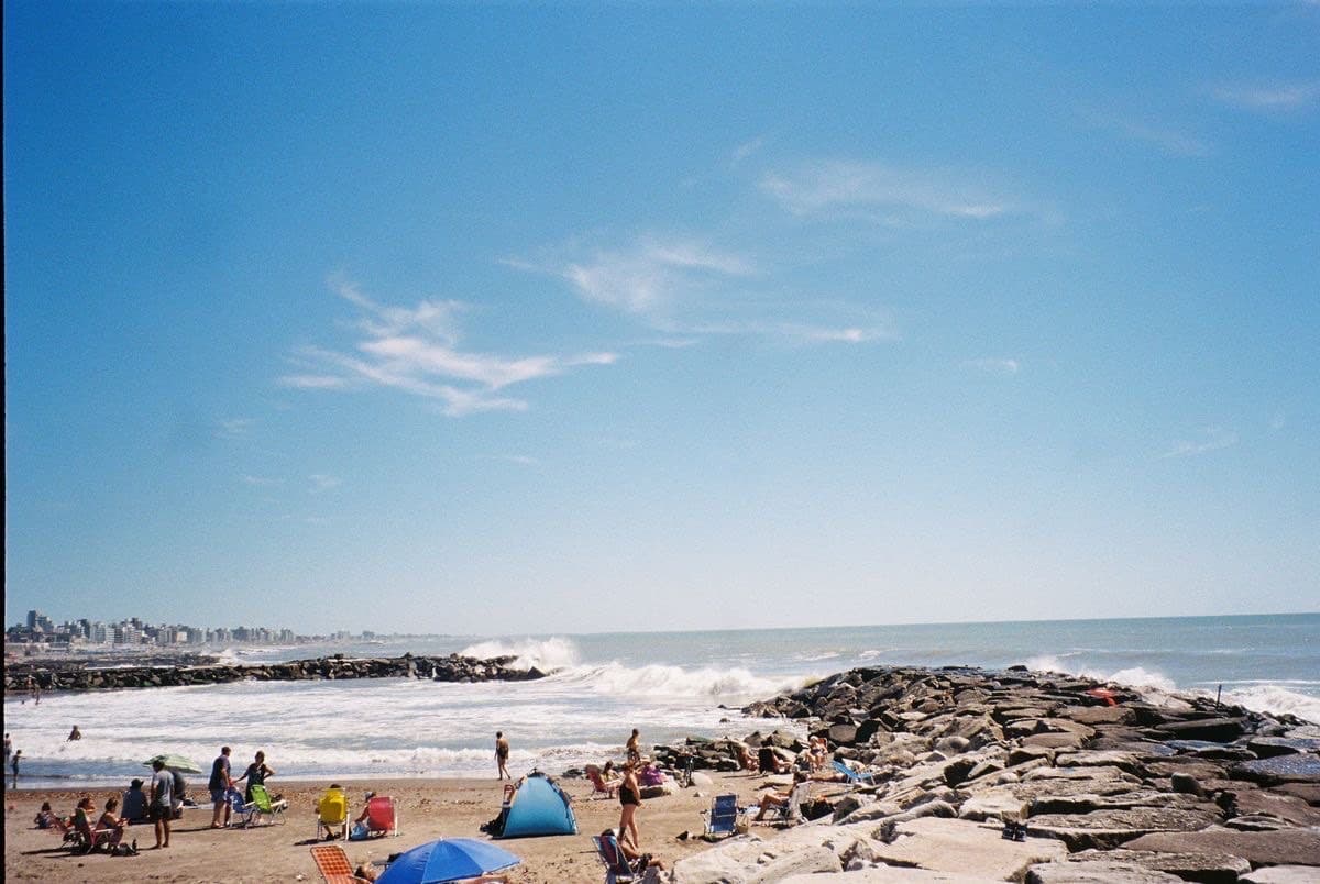Mar del Plata on a summer weekend — sunbathers on the sand, umbrellas, the Atlantic breaking over a rock jetty, the city skyline behind.