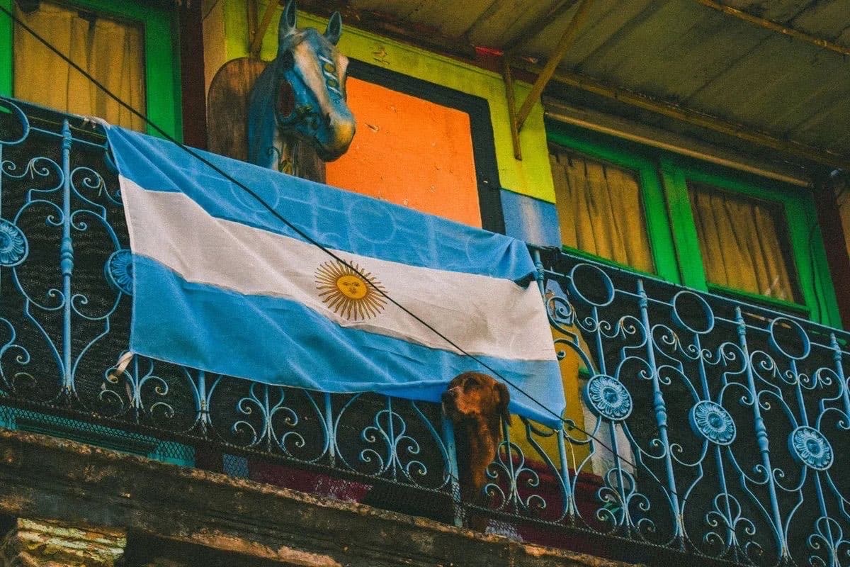 A balcony in La Boca — Argentine flag draped over the wrought-iron railing, wooden horse mounted above, dog watching the street.