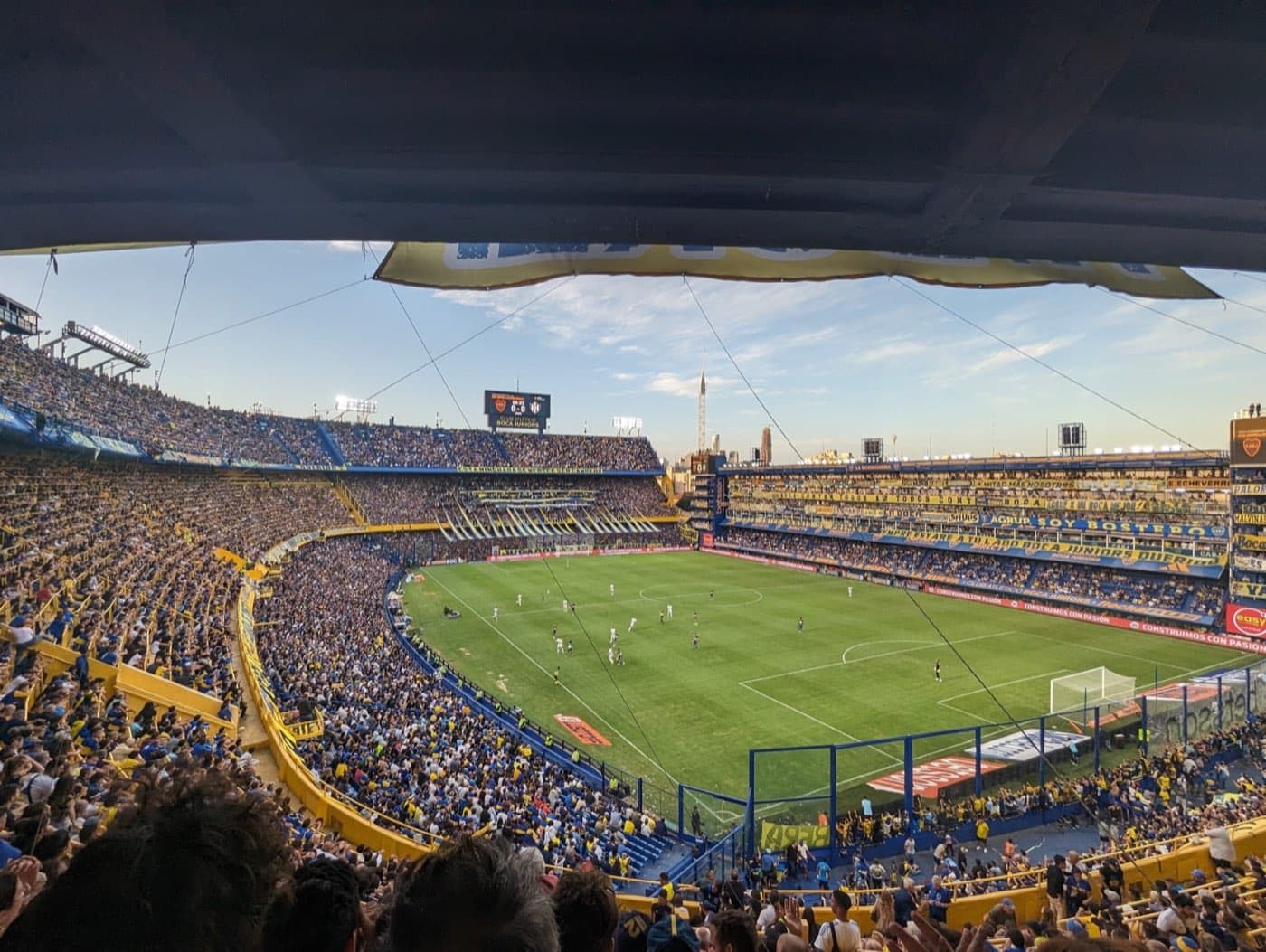 La Bombonera before Boca kick off — packed stand, blue-yellow flags across the terrace, sky pink over Buenos Aires.