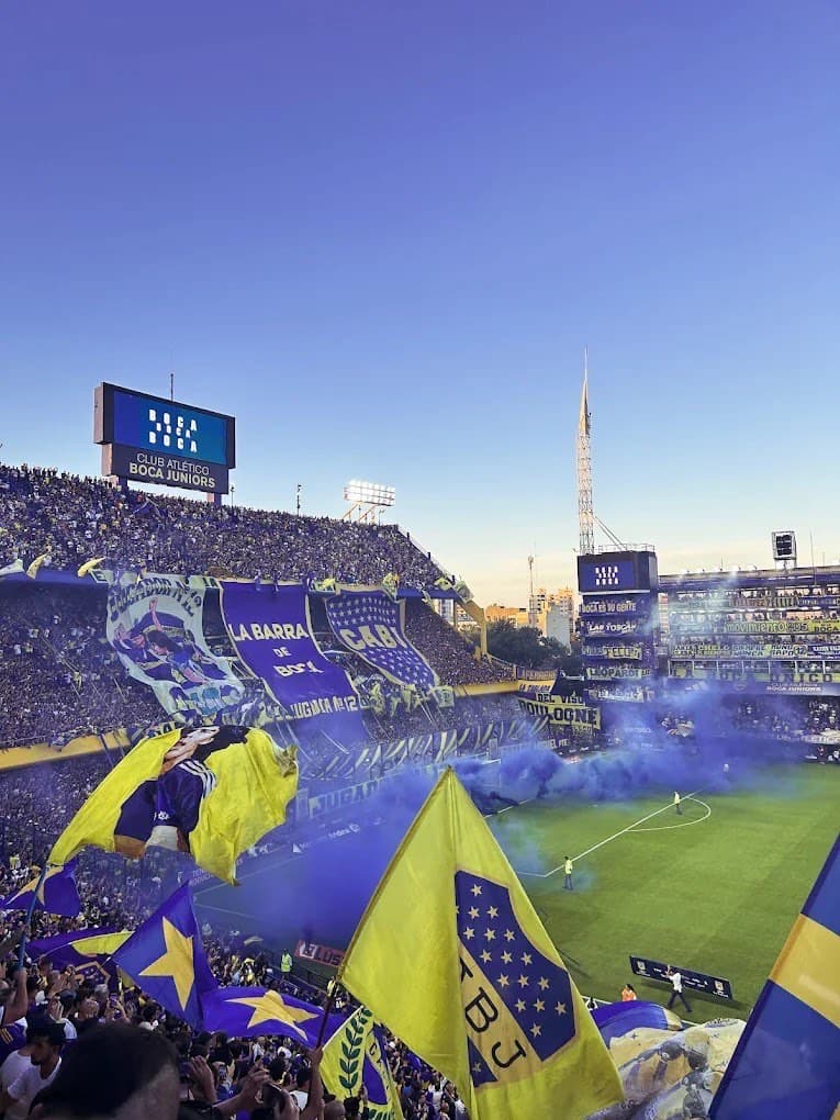 Boca's hinchada in full voice at La Bombonera — blue smoke drifting across the terrace, giant flags, night lights burning through.