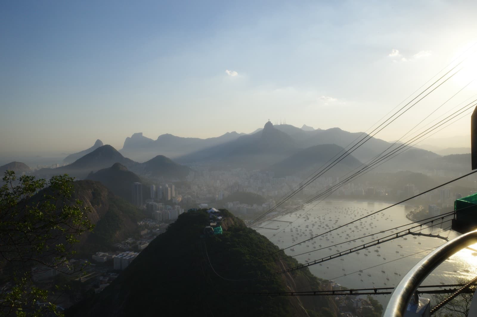 Sugarloaf Mountain at sunset — cable-car cables cutting across the frame, Christ the Redeemer silhouetted on Corcovado in the distance, Botafogo Bay of boats catching the last light.