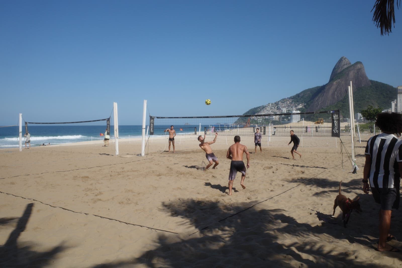 Ipanema beach mid-morning — a footvolley game in play, a Botafogo-striped shirt on the sideline, dog chasing along the sand, Dois Irmãos and the favela climbing the hillside behind.