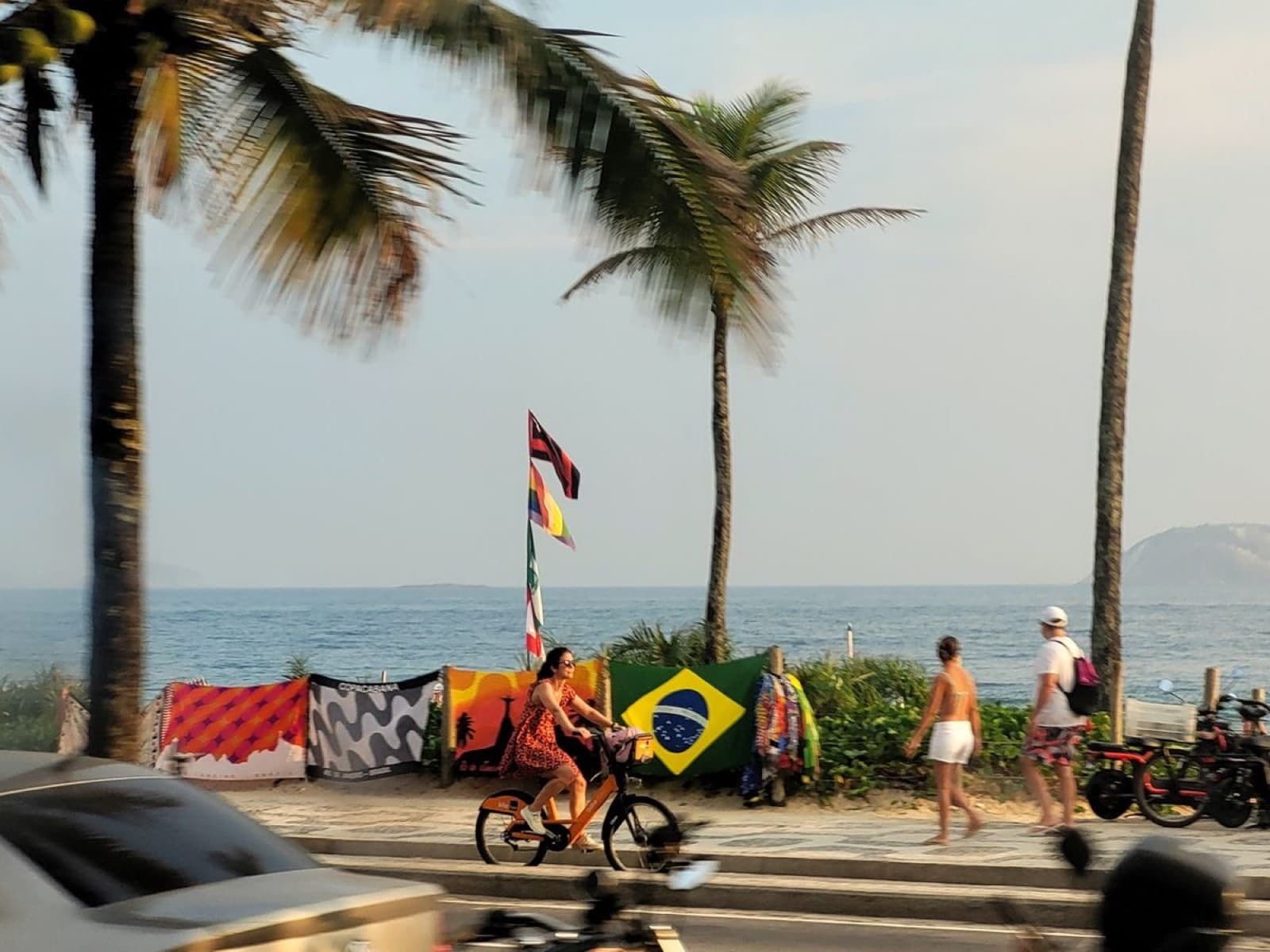 Copacabana promenade at dusk — a woman cycling past palm trees, sarongs and a Brazilian flag hanging on the vendor railing, sea and headland behind.