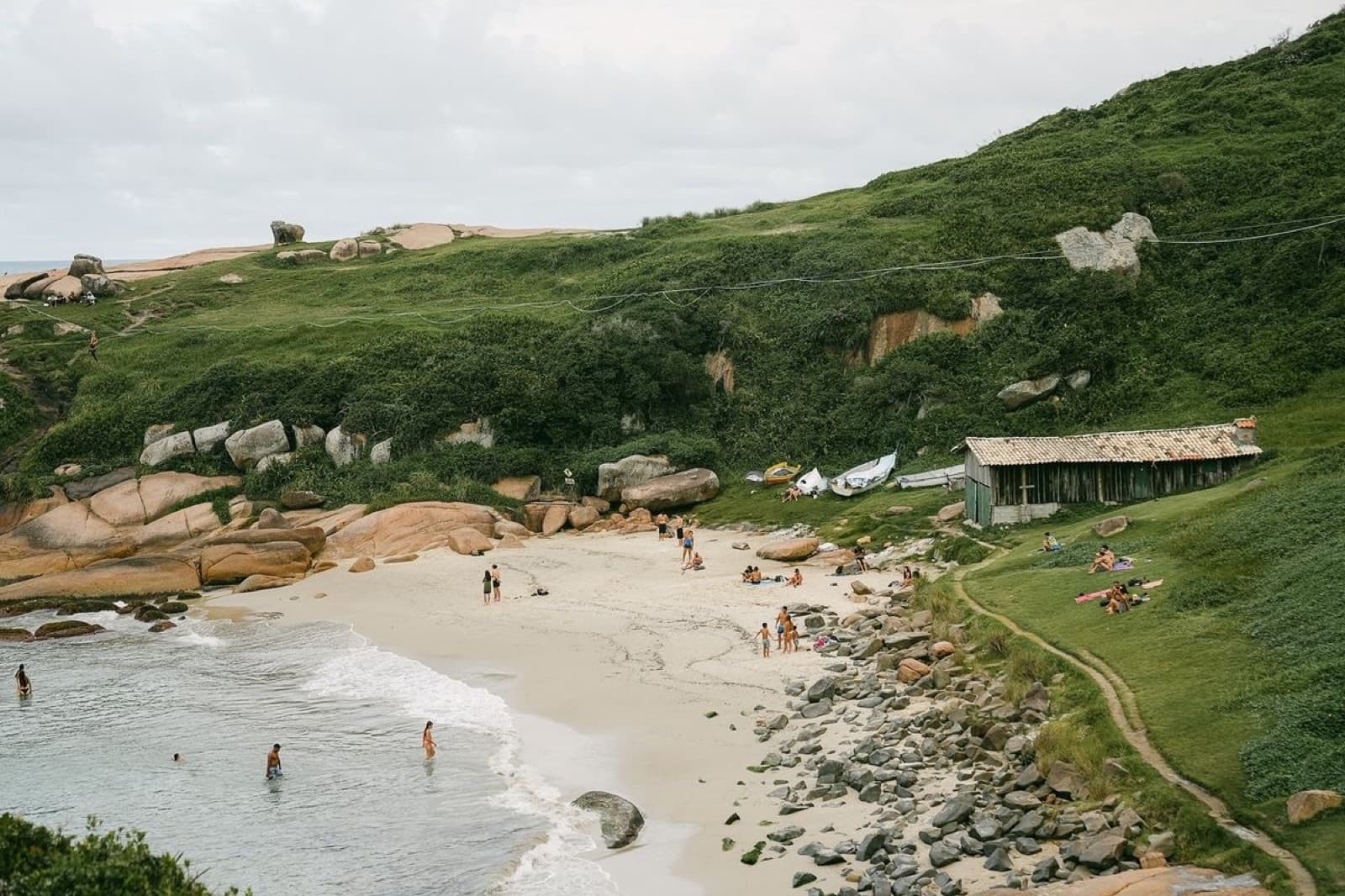 A quiet Florianópolis cove — soft sand between granite boulders, a low green hill behind an old wooden fisherman's shed, swimmers in the shallows.