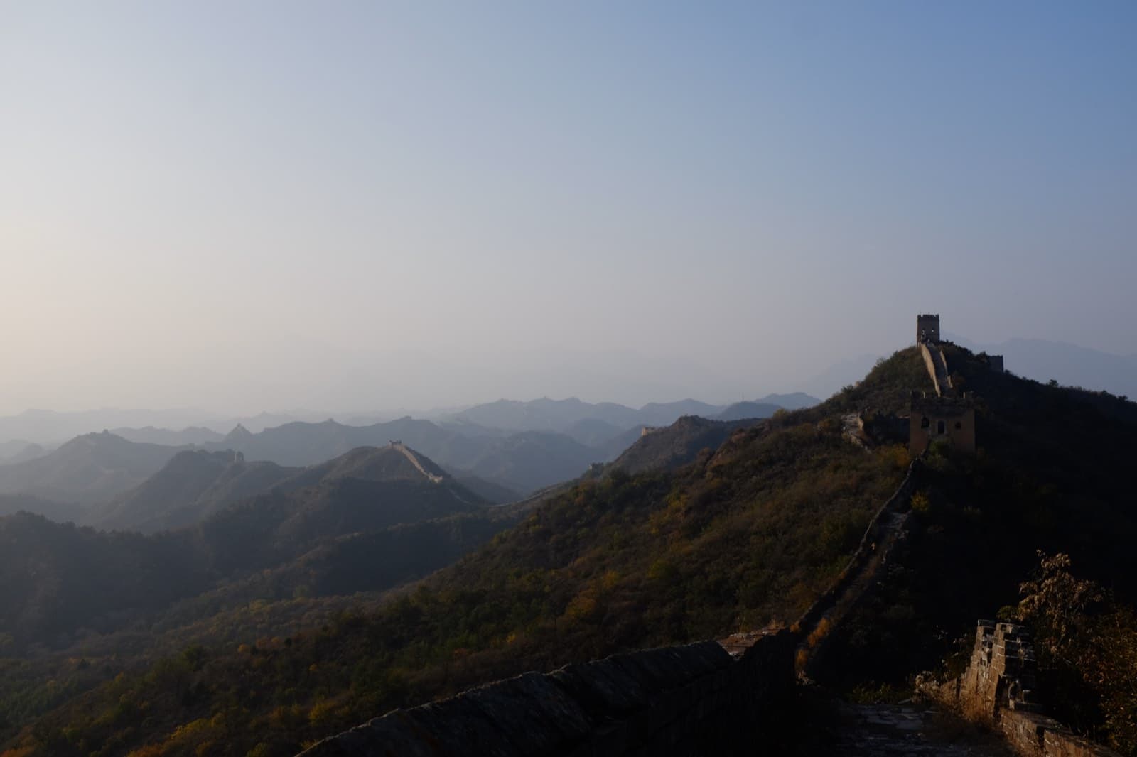 The Great Wall of China at dawn — the wall snaking across layered mountain ridges toward a lone watchtower on the right, the ranges fading into pink-blue haze.