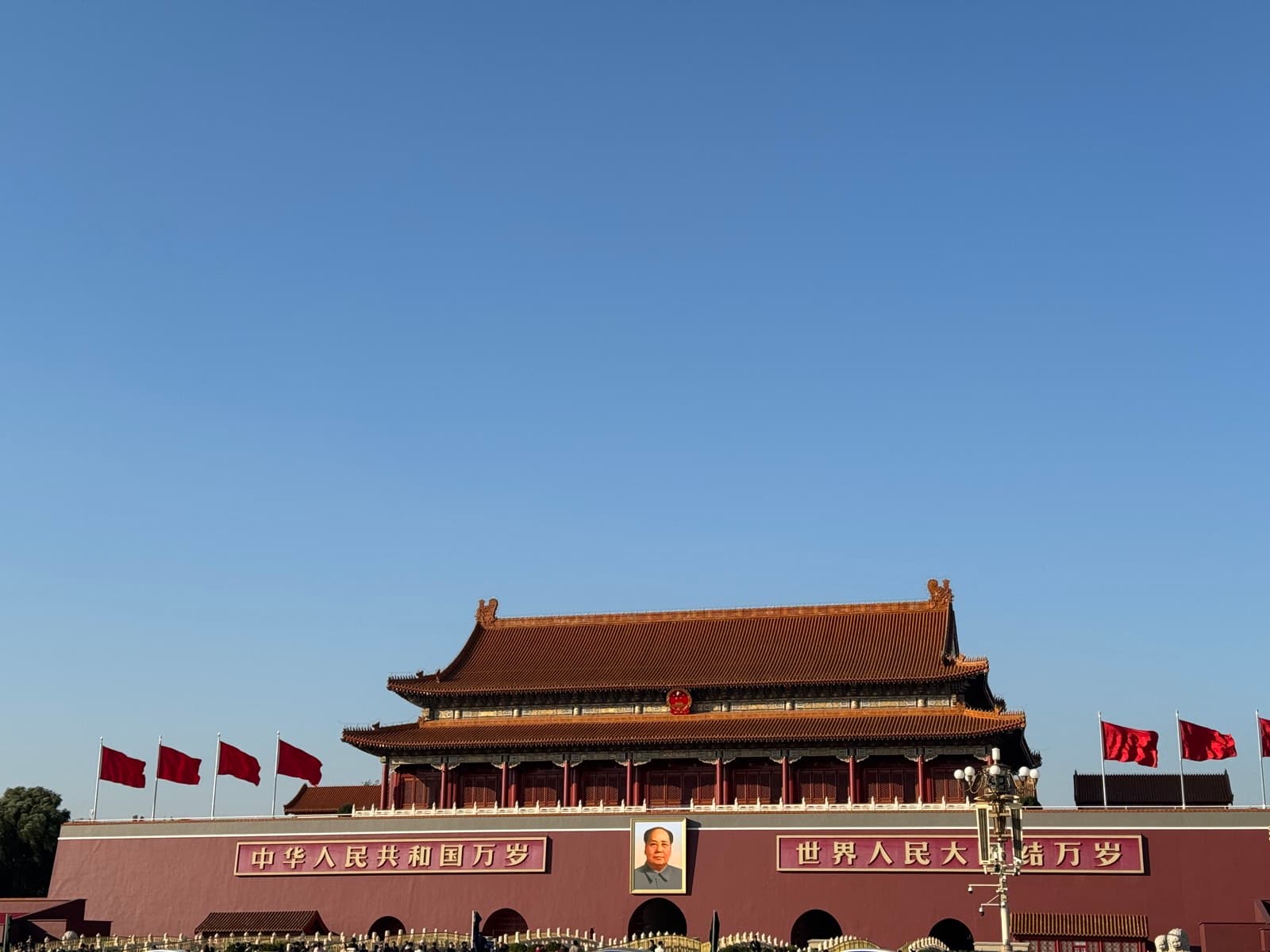 Tiananmen — the Gate of Heavenly Peace with Mao's portrait, red flags flying in a row along the roofline, deep blue Beijing sky above.