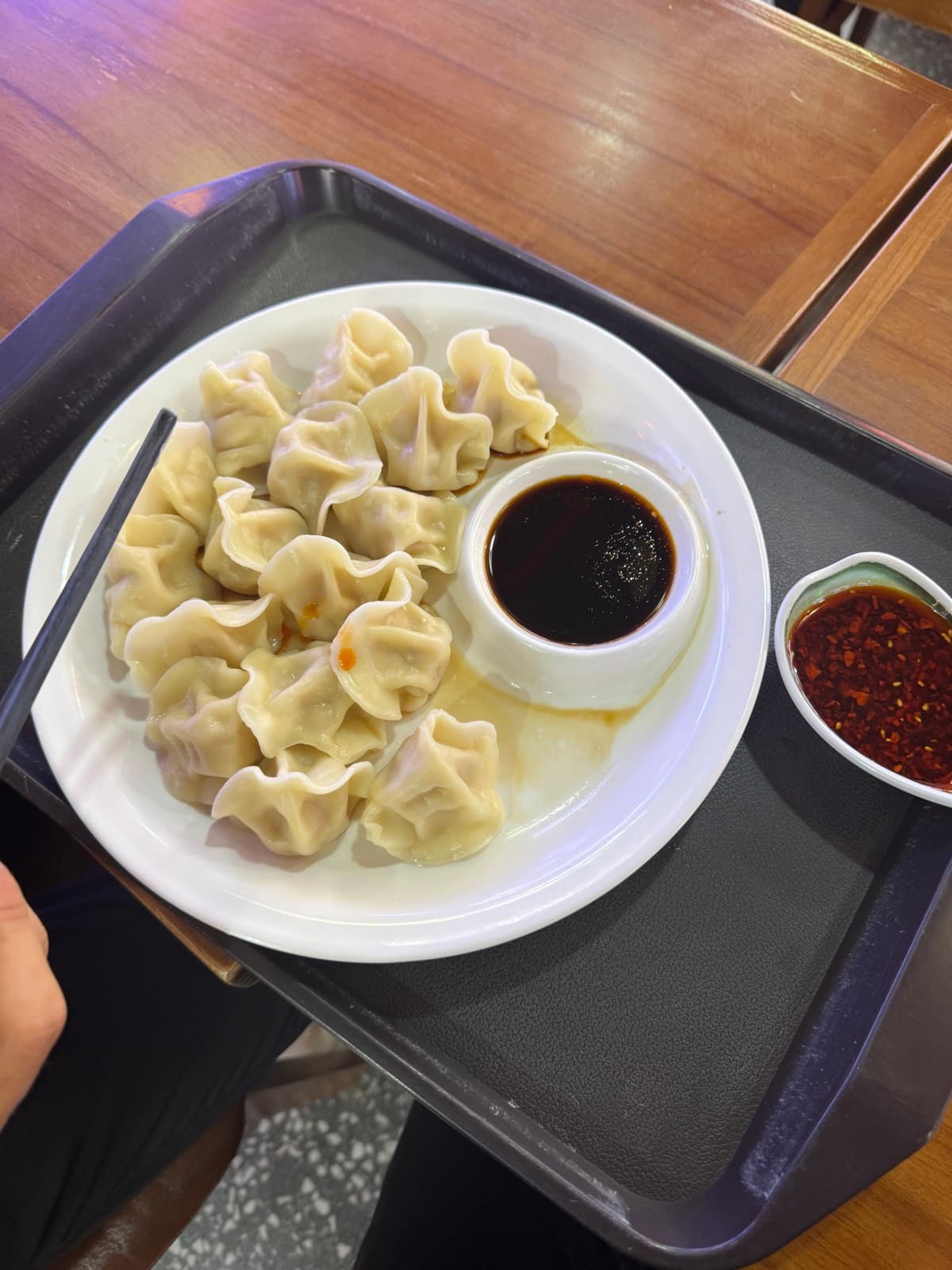 A plate of Chinese jiaozi on a cafeteria tray — soy dip in a ramekin, a side of chilli oil with sesame seeds, chopsticks resting alongside.