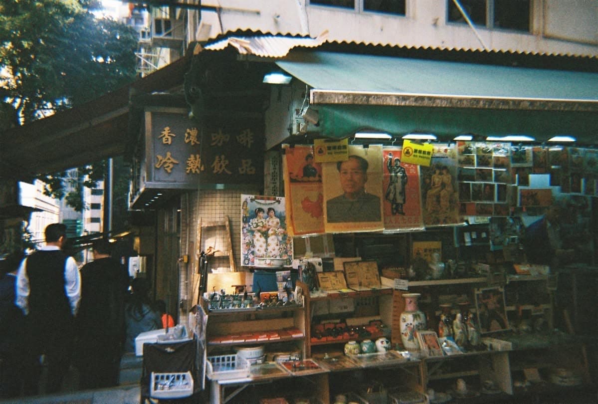 A Hong Kong side-street antique stall at dusk — Mao portraits and Cultural Revolution posters hanging under the awning, a coffee sign in traditional characters above.