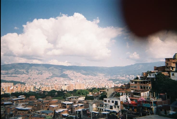 Medellín from a hillside comuna — the city sprawling across the Aburrá Valley under big clouds, mountains rising beyond.