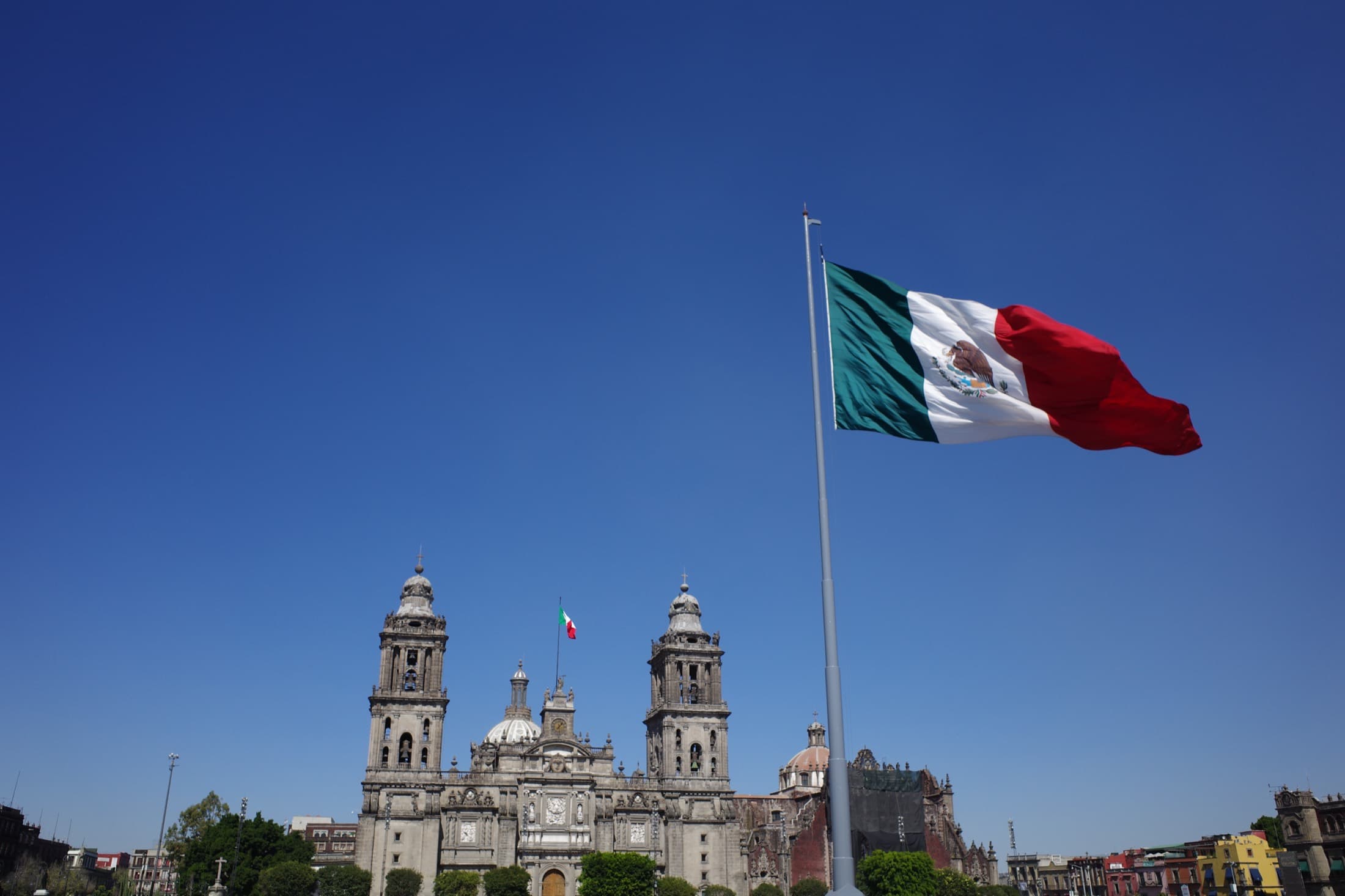 The Metropolitan Cathedral on the Zócalo in Mexico City under a deep-blue sky, the giant Mexican flag billowing from its towering pole over the vast main square.