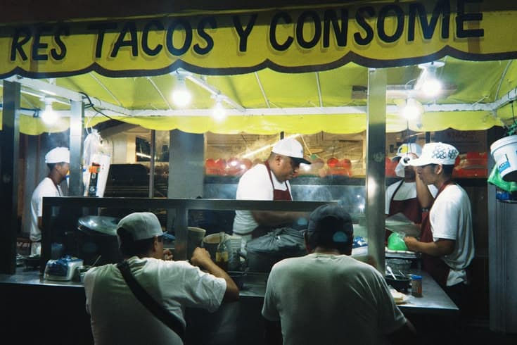 A late-night taco stand under a yellow 'RES TACOS Y CONSOMÉ' awning — cooks in white caps and red aprons working the griddle in the steam while diners lean in at the counter.