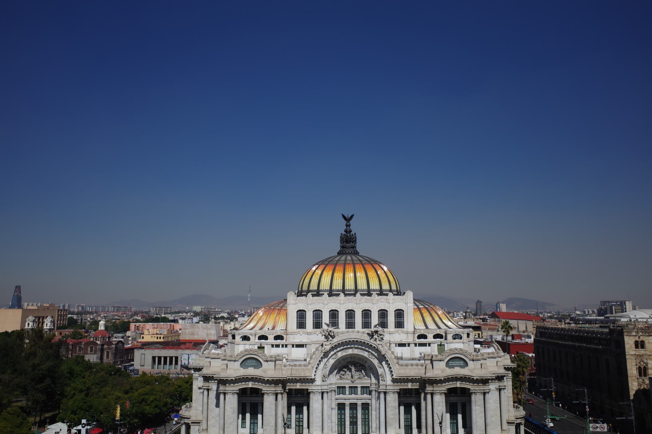 The Palacio de Bellas Artes from above — its white-marble Art Nouveau facade and the famous amber stained-glass dome, the low sprawl of Centro Histórico and the mountains behind.