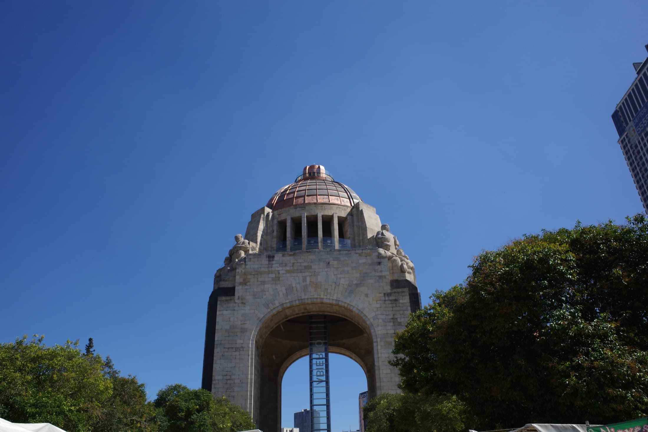 The Monumento a la Revolución from below — the huge stone arch and copper dome with its corner sculpture groups, framed by green trees against a clear blue sky.