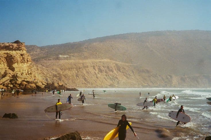 Surfers wading into the break at Imsouane on Morocco's Atlantic coast at golden hour — a dozen boards on the sand and in the shallows below a tall sandstone cliff, one surfer walking out with a yellow board.
