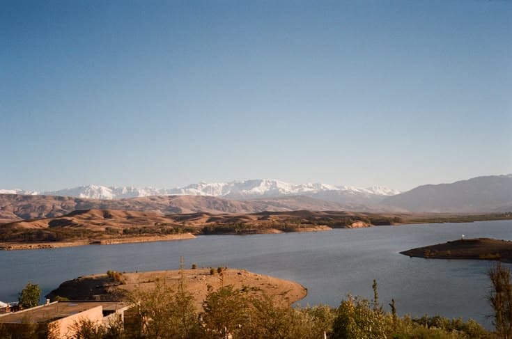 The snow-capped High Atlas mountains rising behind a still reservoir near Marrakech — arid red hills sloping down to the blue water under a clear sky.