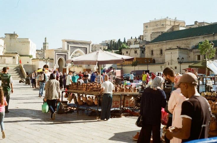 A busy square in the Fez medina — stalls of stacked terracotta pottery and tagines under a parasol, crowds passing an ornately tiled gateway (bab) with the old city climbing the hill behind.