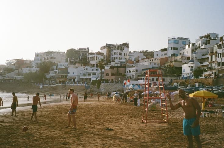 The whitewashed hillside village of Taghazout above its beach at sunset — locals playing football on the sand, a lifeguard tower and yellow umbrellas, the houses stacked up the slope behind.