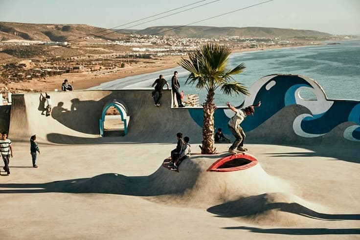 The wave-muralled clifftop skatepark above the coast near Taghazout — kids skating the bowls and a lone palm, the long Atlantic beach and blue-painted wave sculptures stretching to the sea behind.