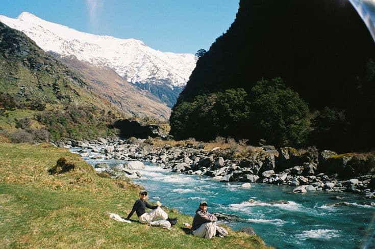 Two trampers resting on the grassy bank of a rushing turquoise braided river deep in a South Island valley, snow-capped peaks glowing at the head of the gorge.
